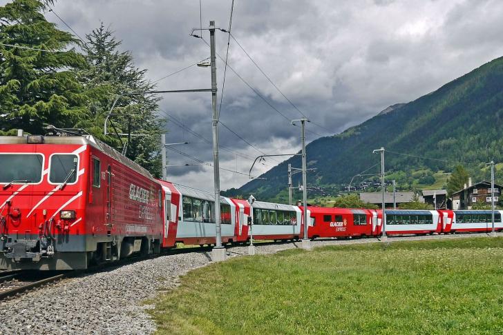 Glacier Express Zug auf Schienen hinter Bergen und grüner Landschaft
